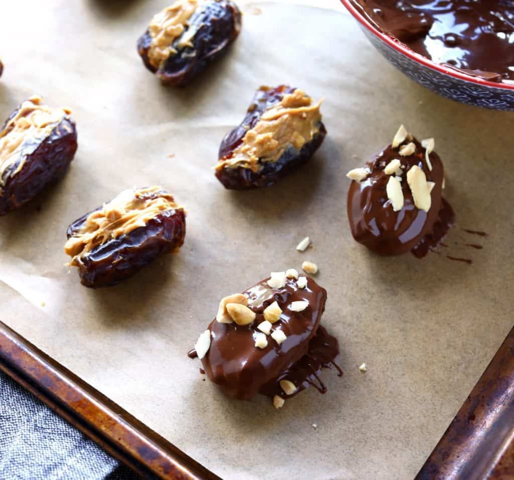 healthy homemade snickers step 2- dip stuffed dates in melted chocolate stuffed with peanut butter and dipped in chocolate on parchment paper ready to freeze #snickerdates #healthysnickers #chocolate #dessert #chocolate #chocolatecovereddates #homemadesnickersrecipe #chocolatecoveredsnickerdates #healthydessertrecipes