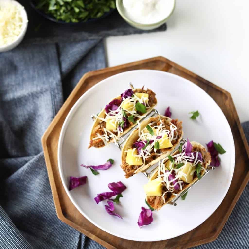 overhead shot of 3 Pineapple pork tacos Al Pastor topped with pinapple, monterey jack, cilantro, red cabbage, and corn tortillas. Served on a white plate a wood platter and a denim napkin.