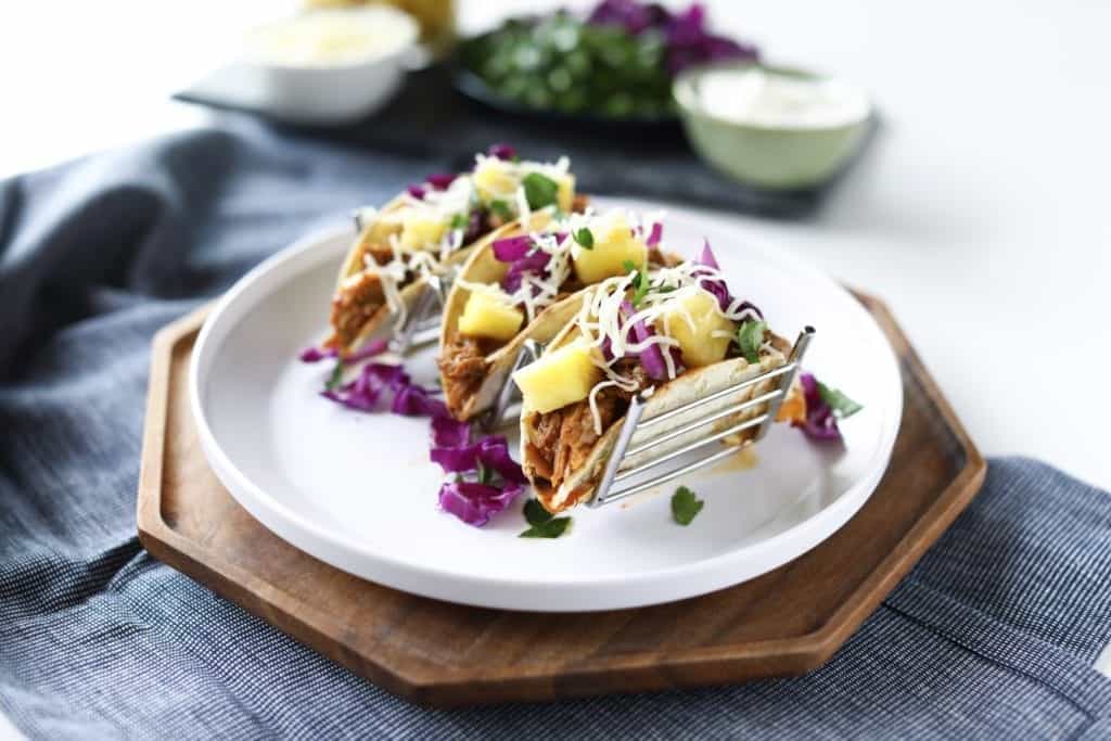 3 Pineapple pork tacos Al Pastor topped with pinapple, Monterey jack, cilantro, red cabbage, and corn tortillas. Served on a white plate a wood platter and a denim napkin. Ready to serve!