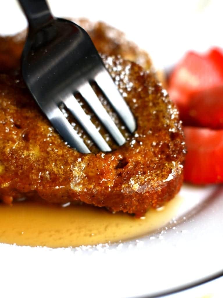 zoomed in picture of pumpkin bread french toast bite with a black fork on a white plate served maple syrup and strawberries. 