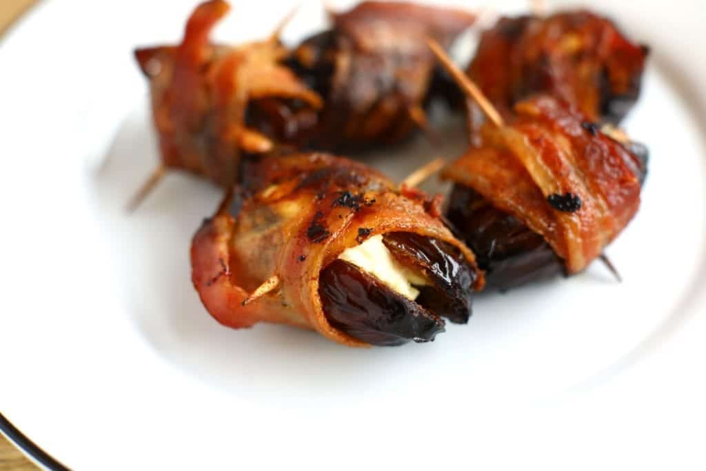 Close up of bacon wrapped dates stuffed with cream cheese on a white plate #bingeworthybites #baconwrappeddateswithcheese #baconwrappeddates #thanksgivingappetizers #appetizers #partyappetizers 
