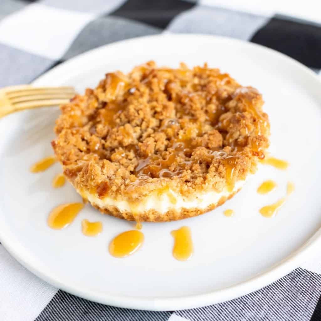 Up close picture of mini caramel apple cheesecakes with crisp streusel topping and caramel drizzle served on white plate/gold fork with black and white plaid table linen #dessert #streusel #cheesecake #caramelapplecheesecake #dessert #streusel #pie #minipies #applepie #baking #easyrecipes #bingeworthybites