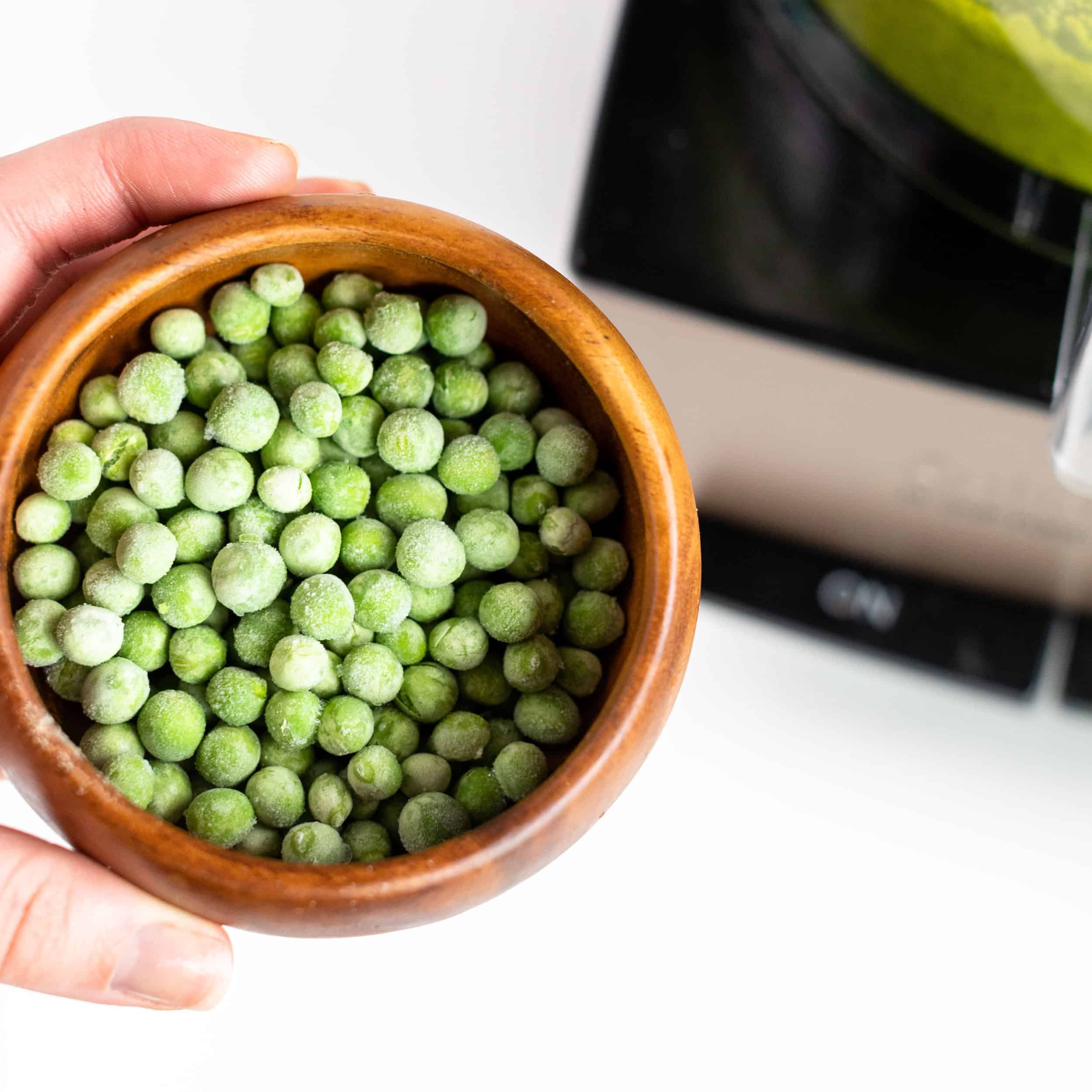 Close up image of frozen peas in a small wooden bowl with food processor in background to add to Fresh Basil Pesto sauce with Toasted Breadcrumbs. Frozen peas will be added to the rest of pesto ingredients. #pestopasta #easyrecipes #fusilli #greenrecipes #weeknightrecipes #pastadish #pasta #breadcrumbs #massimobottura #bingeworthybites