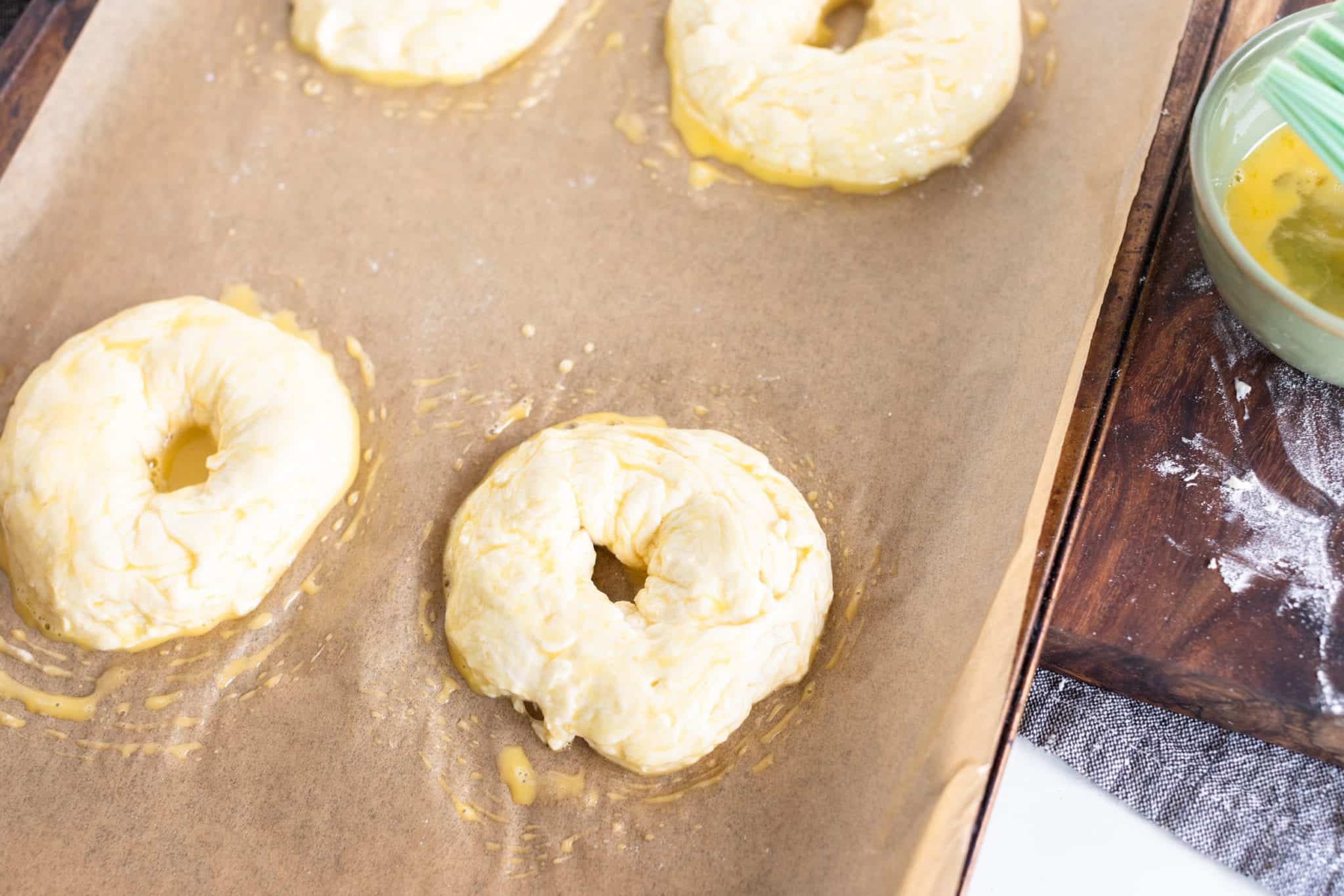 EASY Homemade Everything Bagels with egg wash coating on lined baking sheet before they get seasoned. EASY Homemade Everything Bagels
